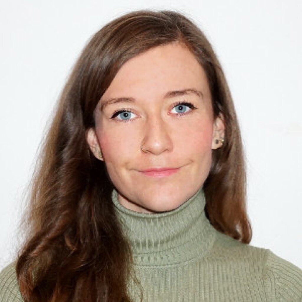 Teresa lächelt in die Kamera, trägt Ohrringe und einen grünen Rollkragen-Pullover. Sie hat langes braunes Haar.